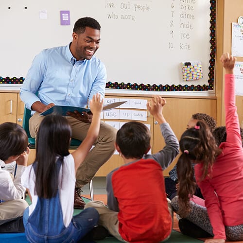 Young children sitting down in front of teacher who is holding an open book as they raise their hand 