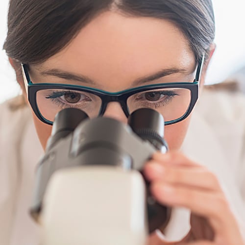  Young female technician looking into microscope in medical laboratory