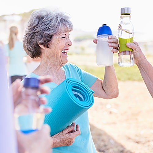 Two elderly women celebrating the end of their yoga class by tapping their water jugs and holding their mats 