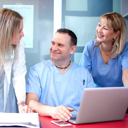 Two nurses smiling at a doctor while one of them sits in front of a computer