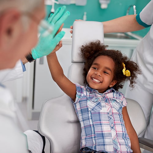 Child on treatment chair high-fiving dentist as dental hygienist stands there
