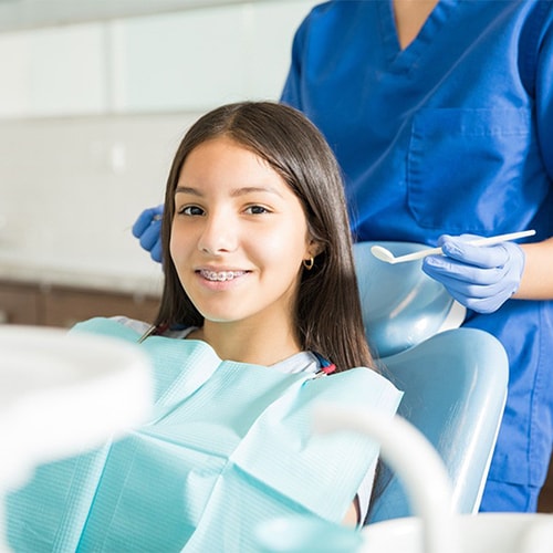 A young female teen with braces is sitting in a treatment chair  smiling at the camera while the dental hygienist stands behind her