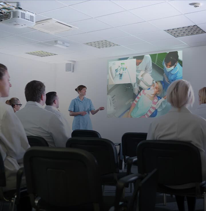 A classroom of dental professionals wearing white coats looking at a projected presentation